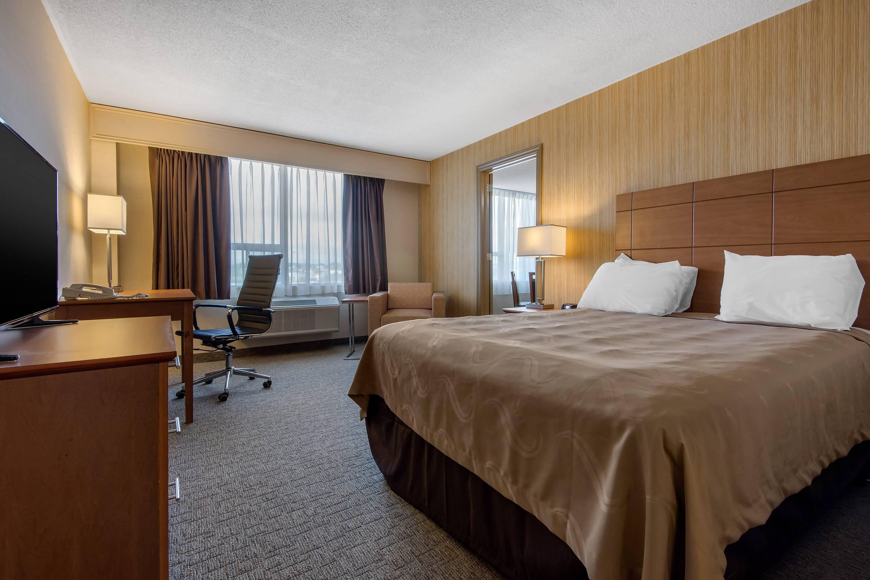 A hotel room with a large bed, a desk with a chair, a TV, and a window with curtains.