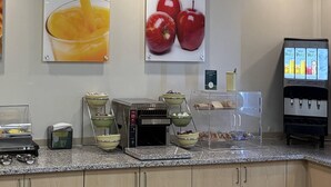 Una barra de desayuno con una máquina de café, cuencos de fruta y dispensador de jugo.
