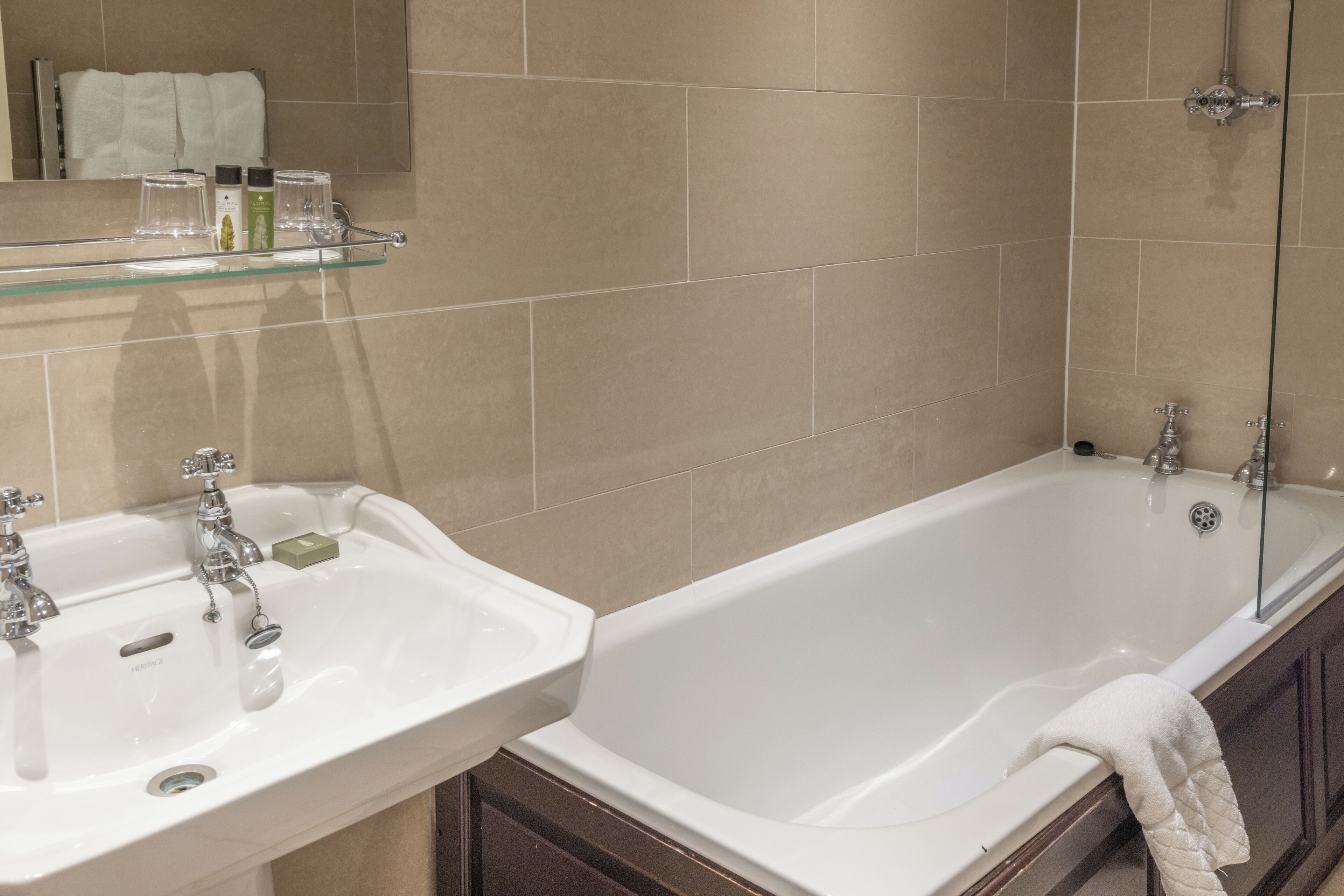 Un baño con lavamanos doble, bañera y estante de vidrio con artículos de aseo.
