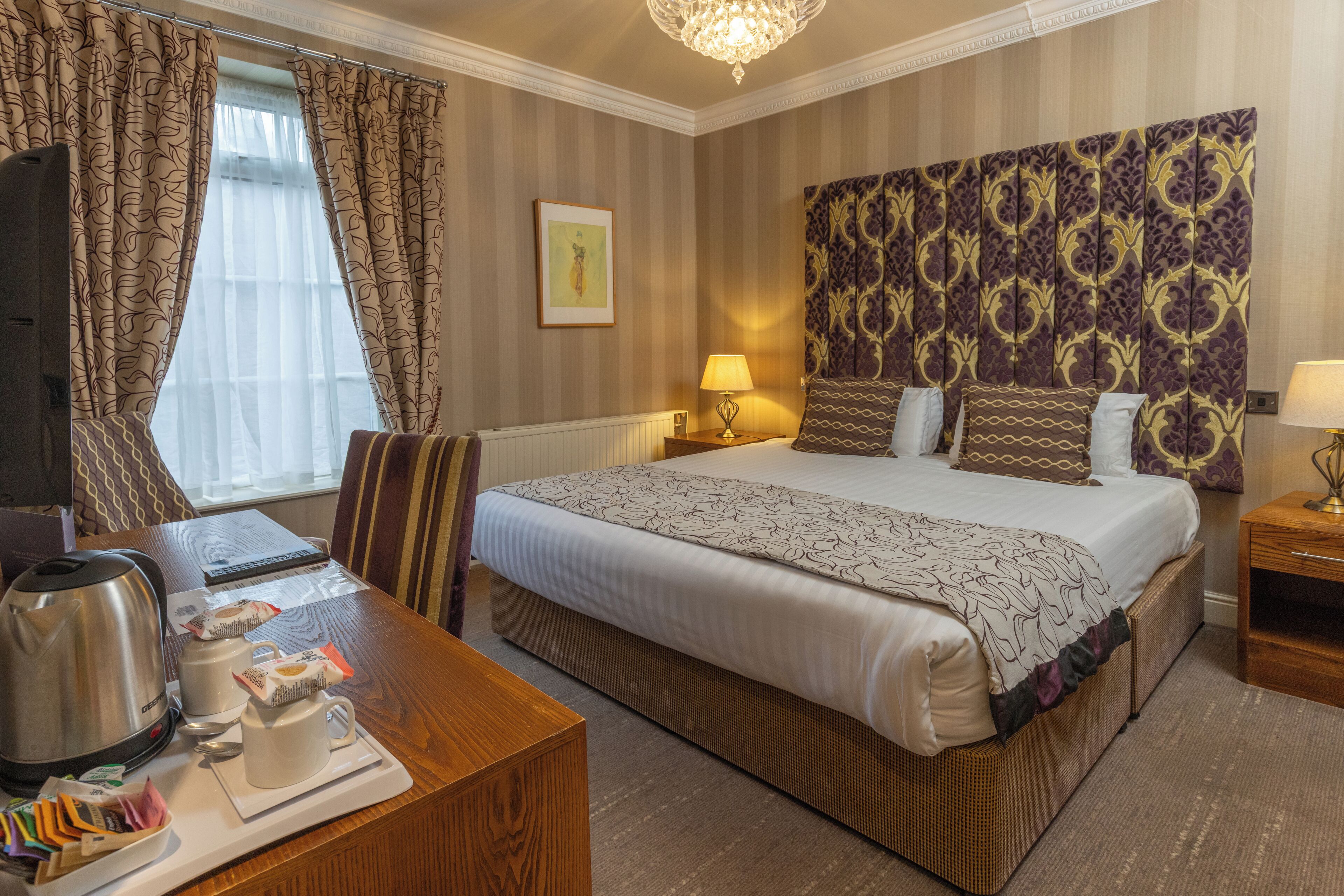 A hotel room with a bed, bedside tables, a desk with a kettle, and a television.