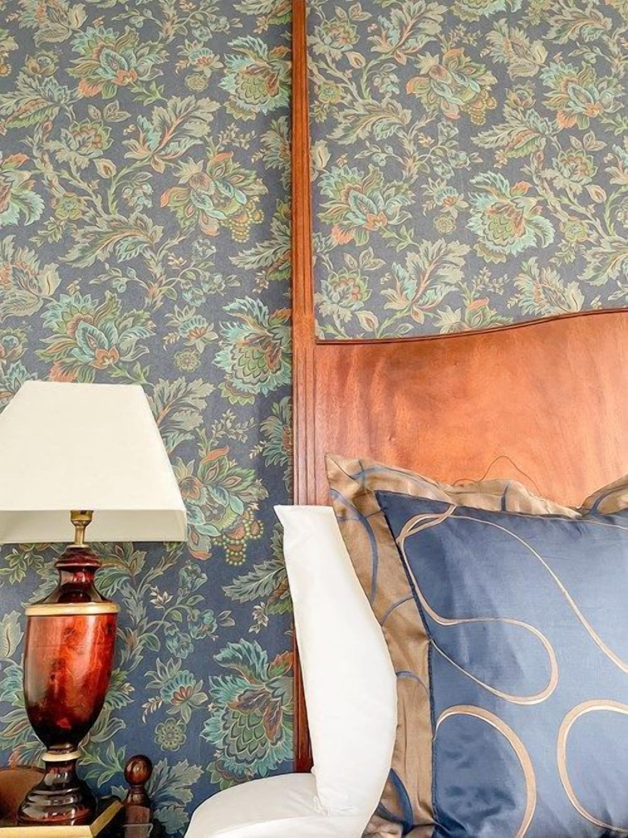 A room with floral wallpaper, a wooden headboard, a white chair, and a table with a lamp.