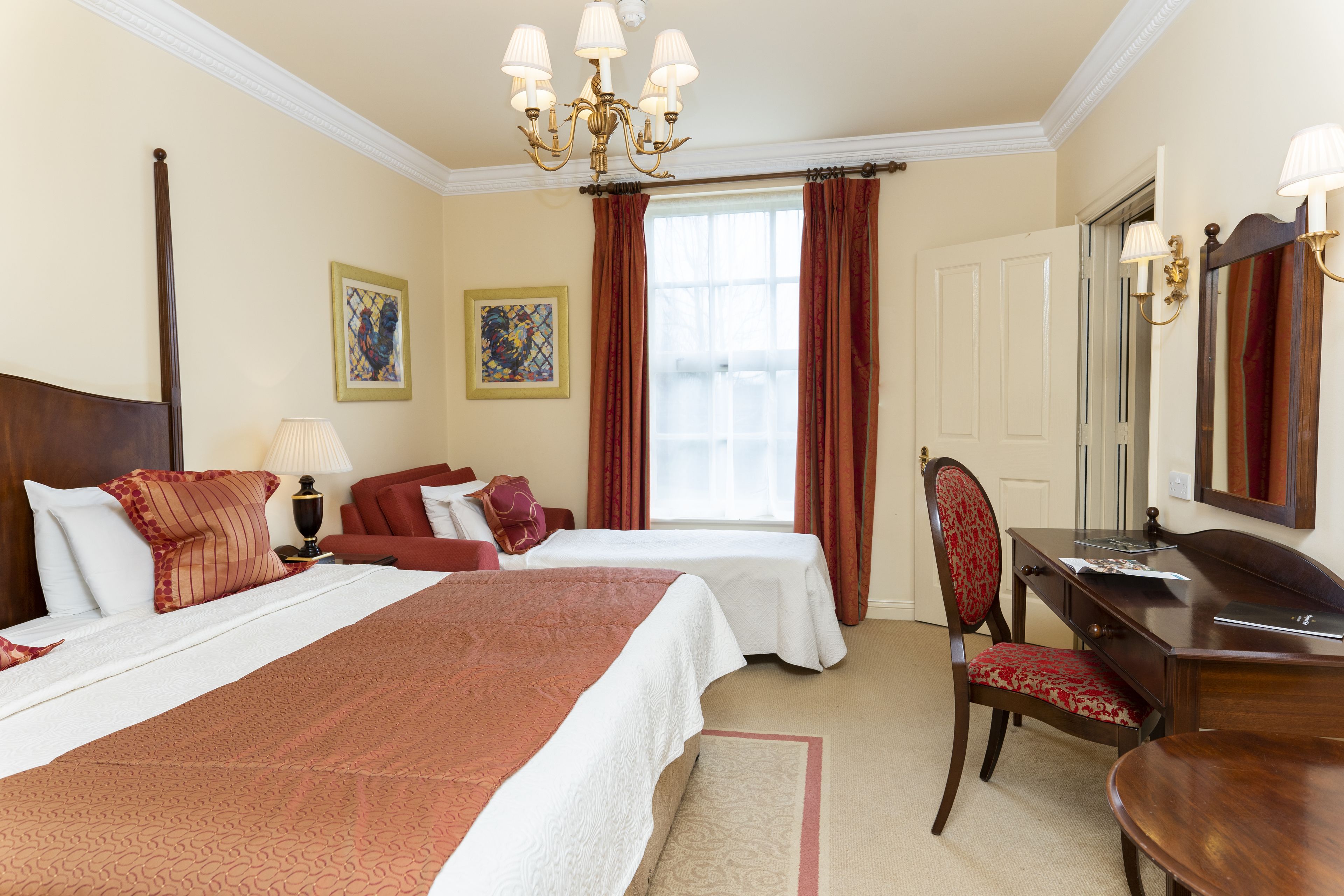 A hotel room with two beds, a desk, a chair, a desk lamp, a chandelier, and a window with curtains.