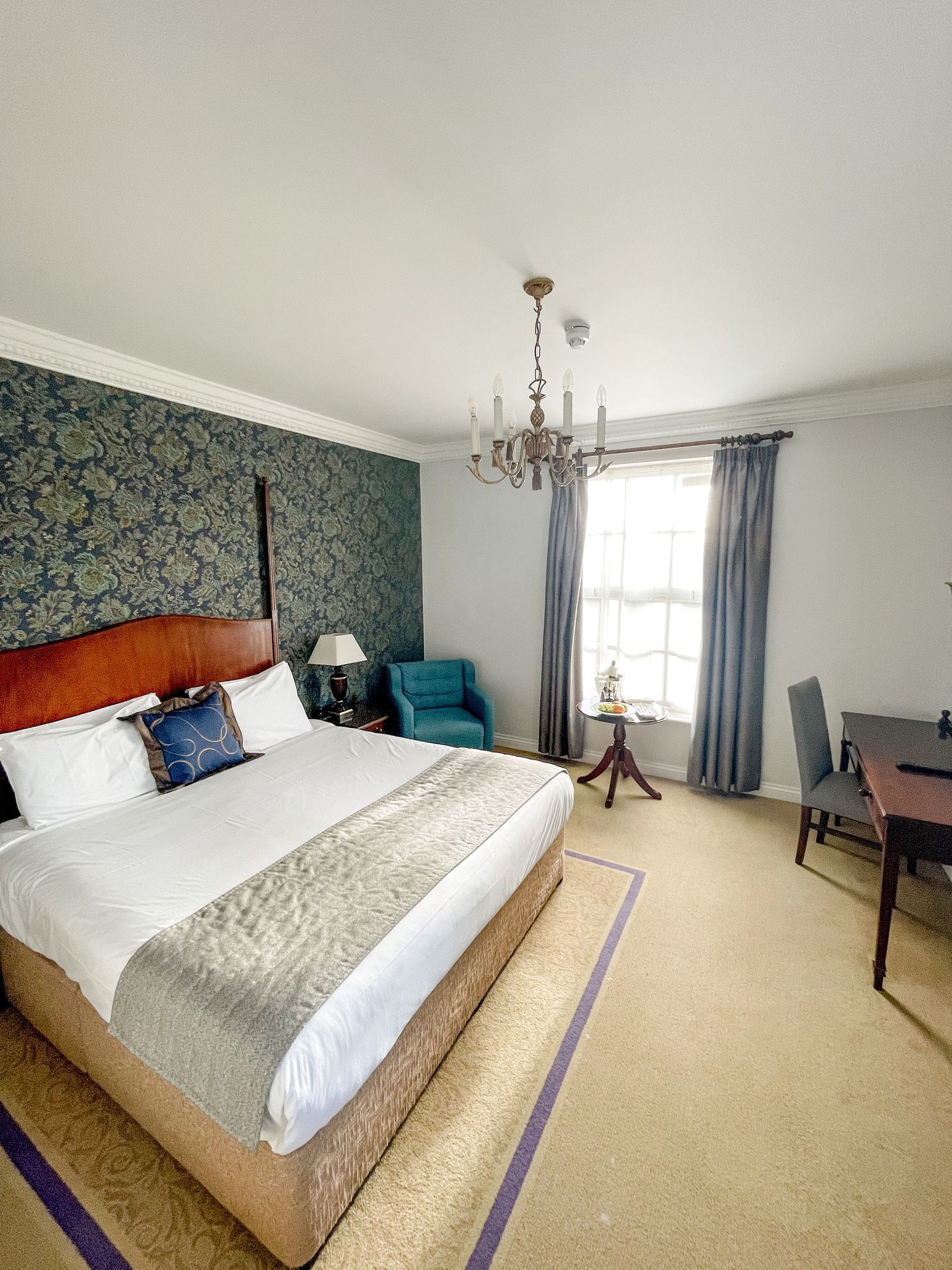 A hotel room with a large bed, a desk, a chair, a chandelier, and a window with curtains.