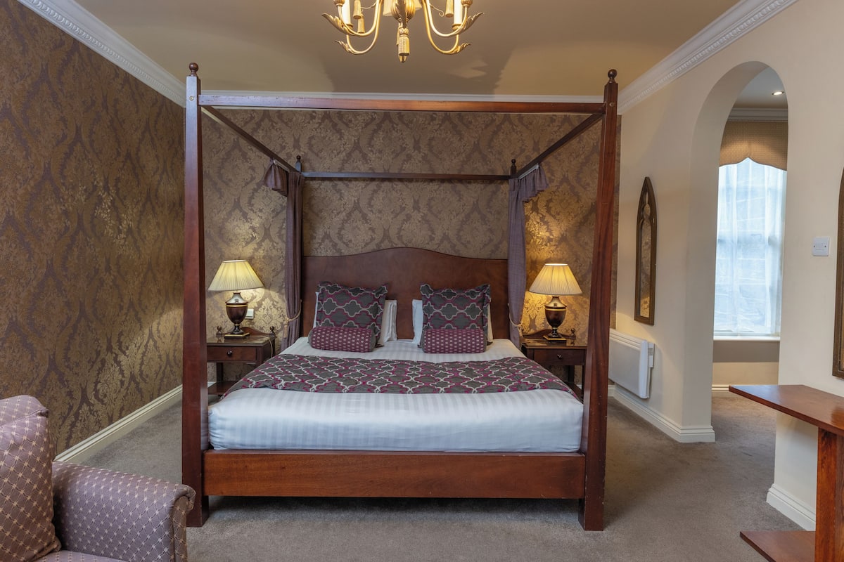 A four-poster bed with a wooden frame, two bedside tables with lamps, patterned wallpaper, and a chandelier.