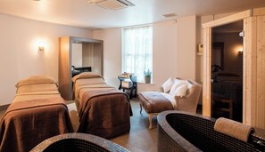 A spa room with two massage tables, a small desk with a computer, a window with sheer curtains, and a wooden door leading to another room. - Ringwood Hall Hotel & Spa, BW Signature Collection (Chesterfield)