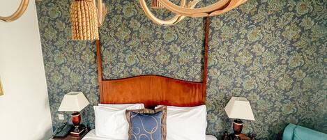 A large bed with a wooden headboard, two bedside tables with lamps, a blue armchair, and a chandelier.