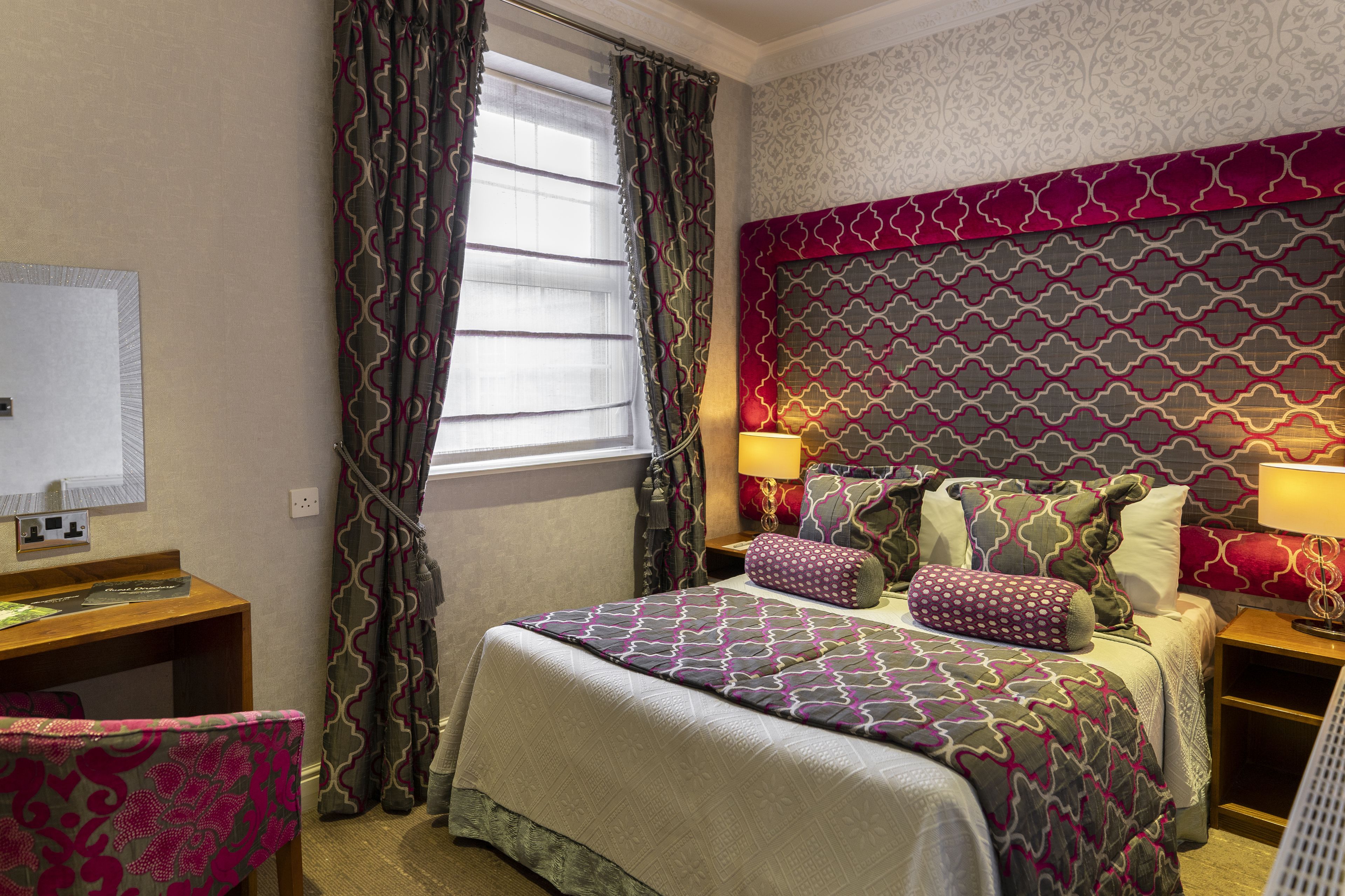 Habitación de hotel con cama, escritorio y silla. Se ve una ventana con cortinas, una pared con papel tapiz estampado y una mesita de noche con lámpara.