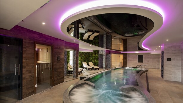 Et moderne poolområde indendørs med jacuzzi, liggestole og en øvre etage med glasfacade.
