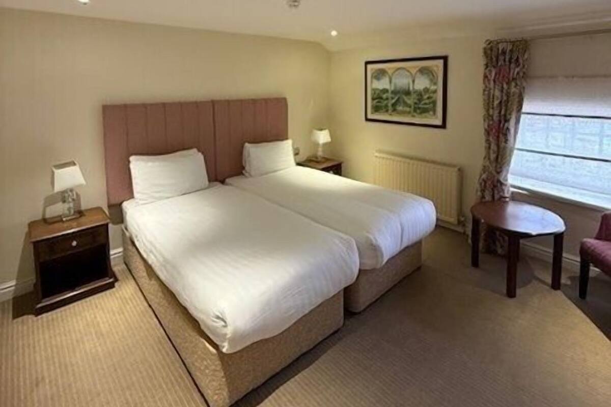 A hotel room with a double bed, bedside tables with lamps, a small round table, a chair, a radiator, and a window with curtains.