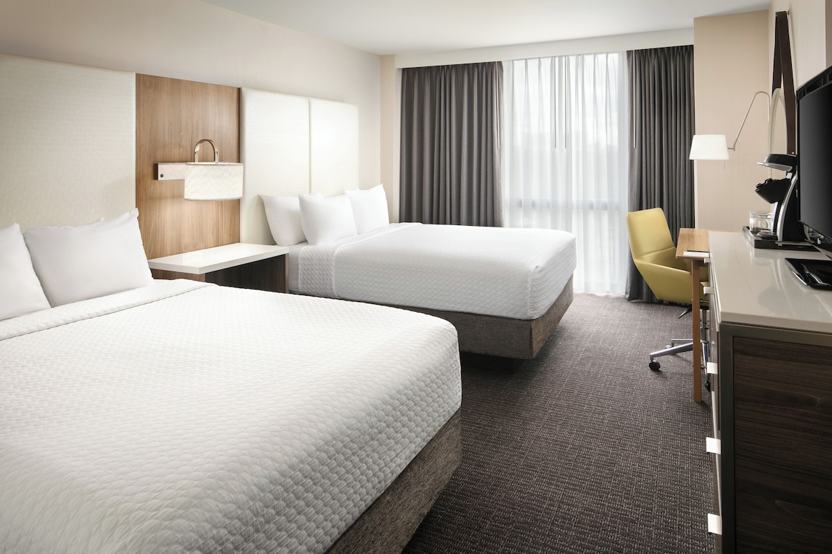 A hotel room with two beds, a desk, a chair, a TV, and a window with curtains.