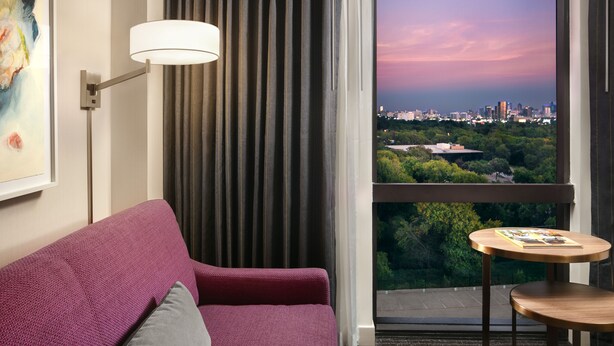 A modern hotel room with a purple sofa, a small round table, a lamp, and a view of a cityscape at dusk. - Crowne Plaza Dallas Market Ctr - Love Field by IHG (Dallas)