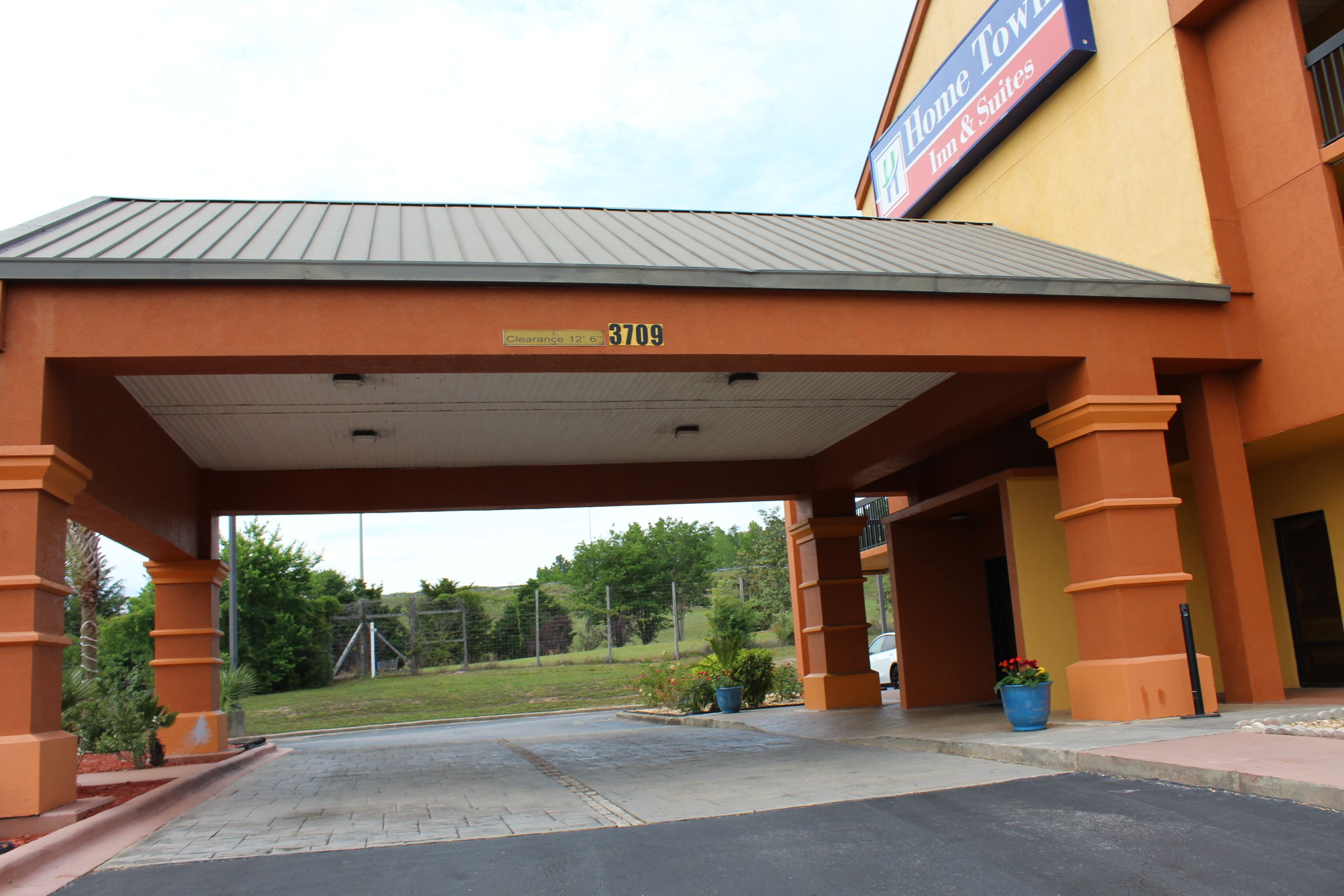 A hotel entrance with a covered driveway, a sign indicating 'Home Town Inn & Suites', and a clear view of the surrounding greenery.