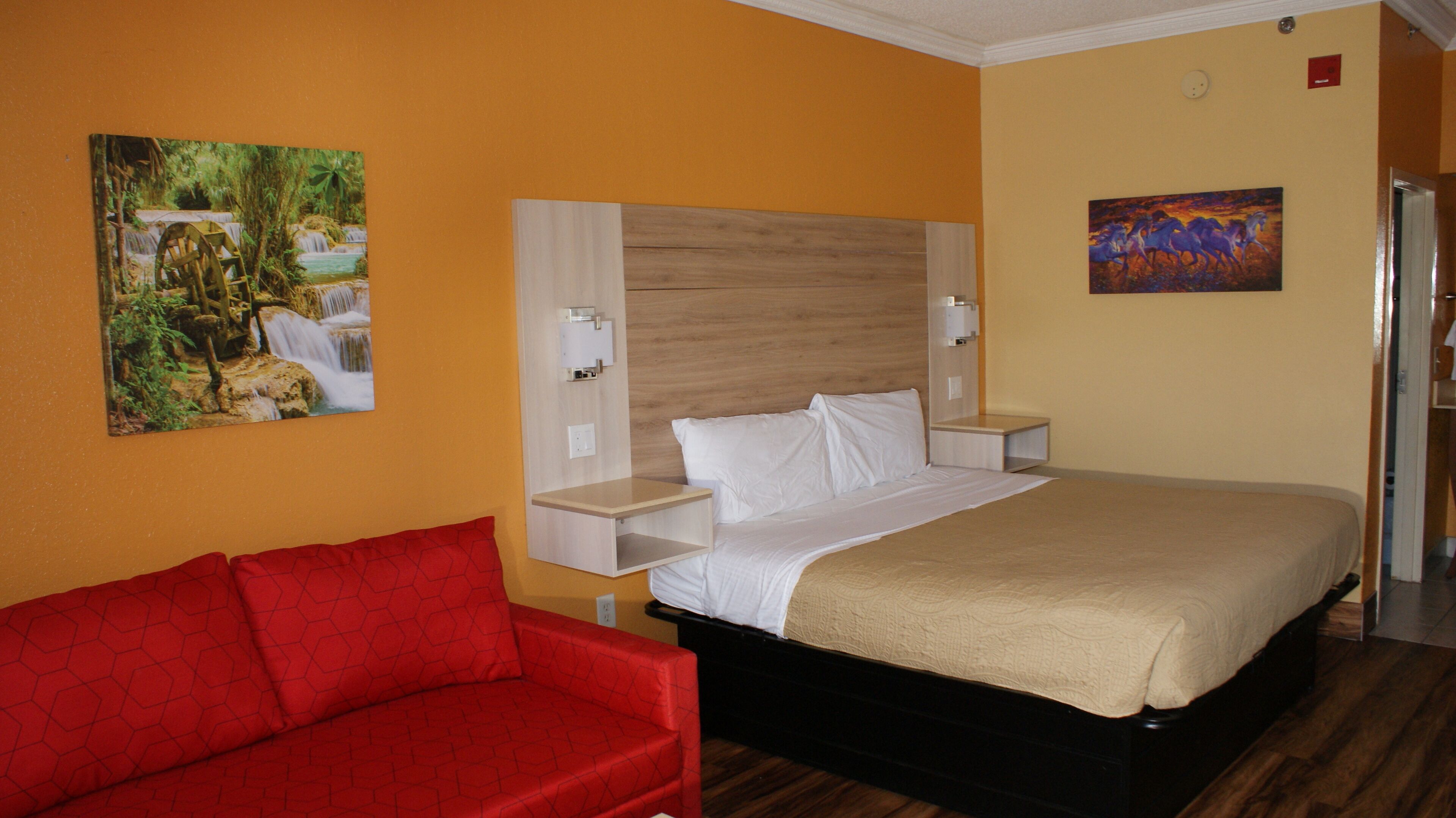 Habitación de hotel con cama, sofá rojo, cabecero de madera, dos mesitas de noche y dos lámparas de pared.