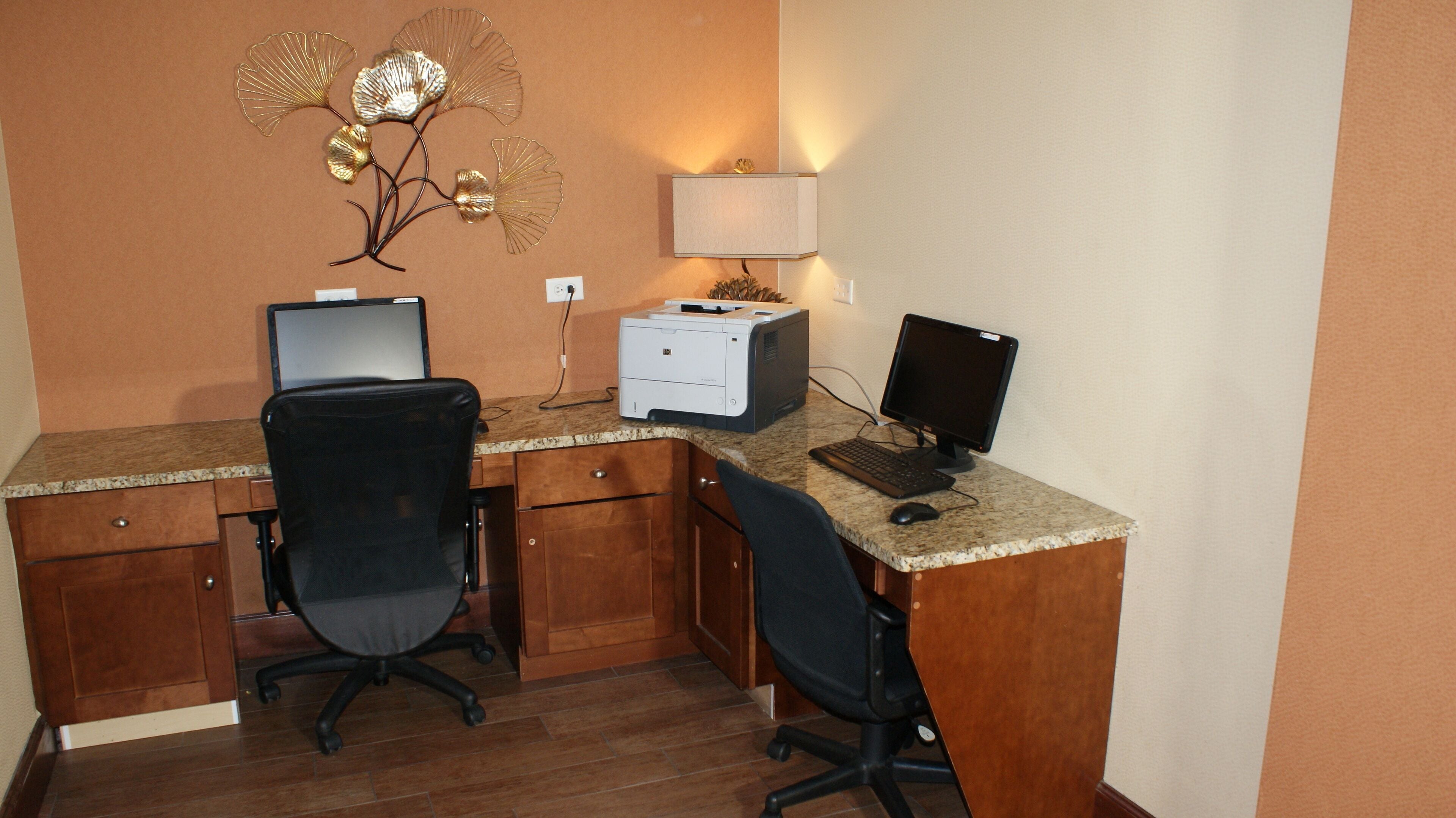 A small office with two desks, each with a computer and chair. There is a printer and a lamp on the wall.