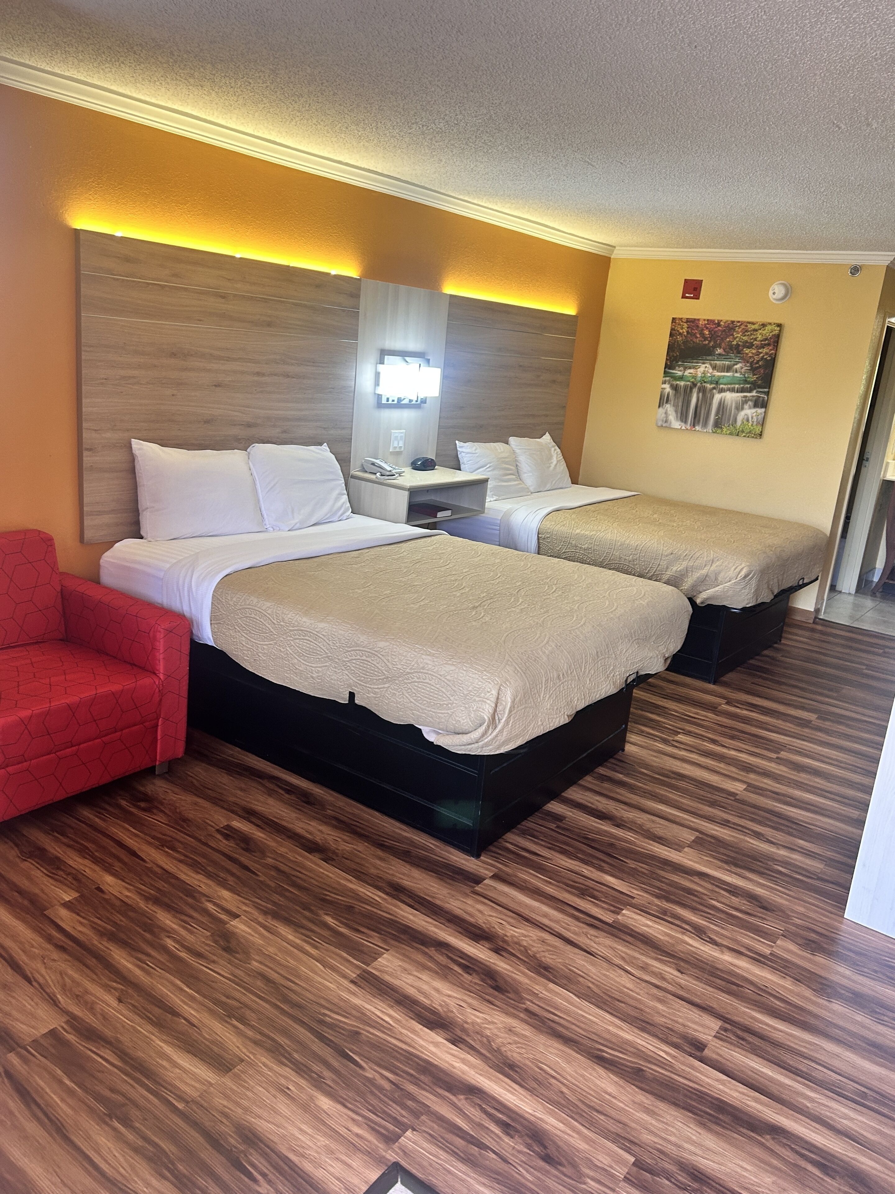 A hotel room with two beds, a red chair, a nightstand with a phone, and a wall-mounted light.