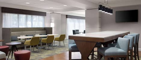 Una habitación de hotel moderna con sofá, mesas redondas, sillas amarillas, una mesa de comedor de madera y un televisor montado en la pared.