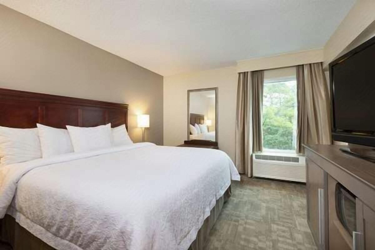 Quarto de hotel com cama grande, televisão de tela plana, espelho e janela com cortinas.