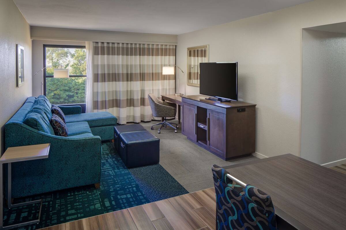 Una habitación de hotel moderna con un sofá modular, un escritorio con silla, un televisor de pantalla plana y un gran ventanal con cortinas.