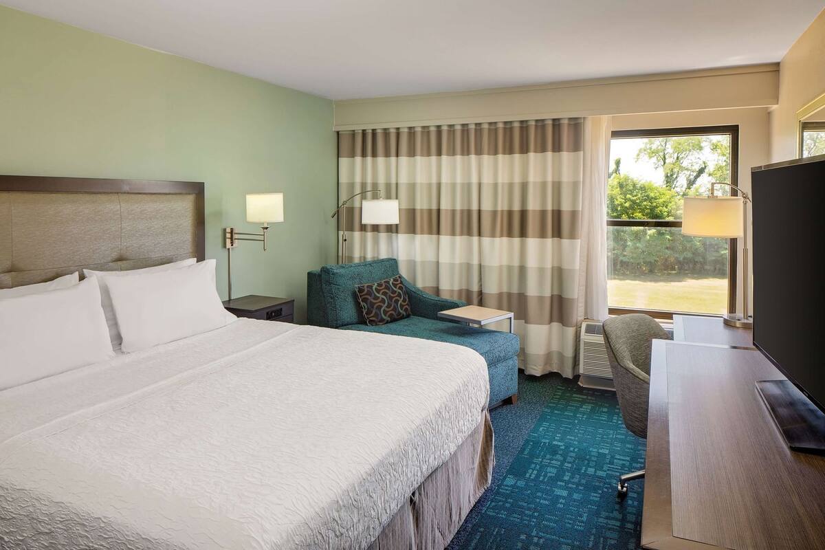 Habitación de hotel con una cama grande, un escritorio, una silla, un televisor, una lámpara y una ventana con cortinas.