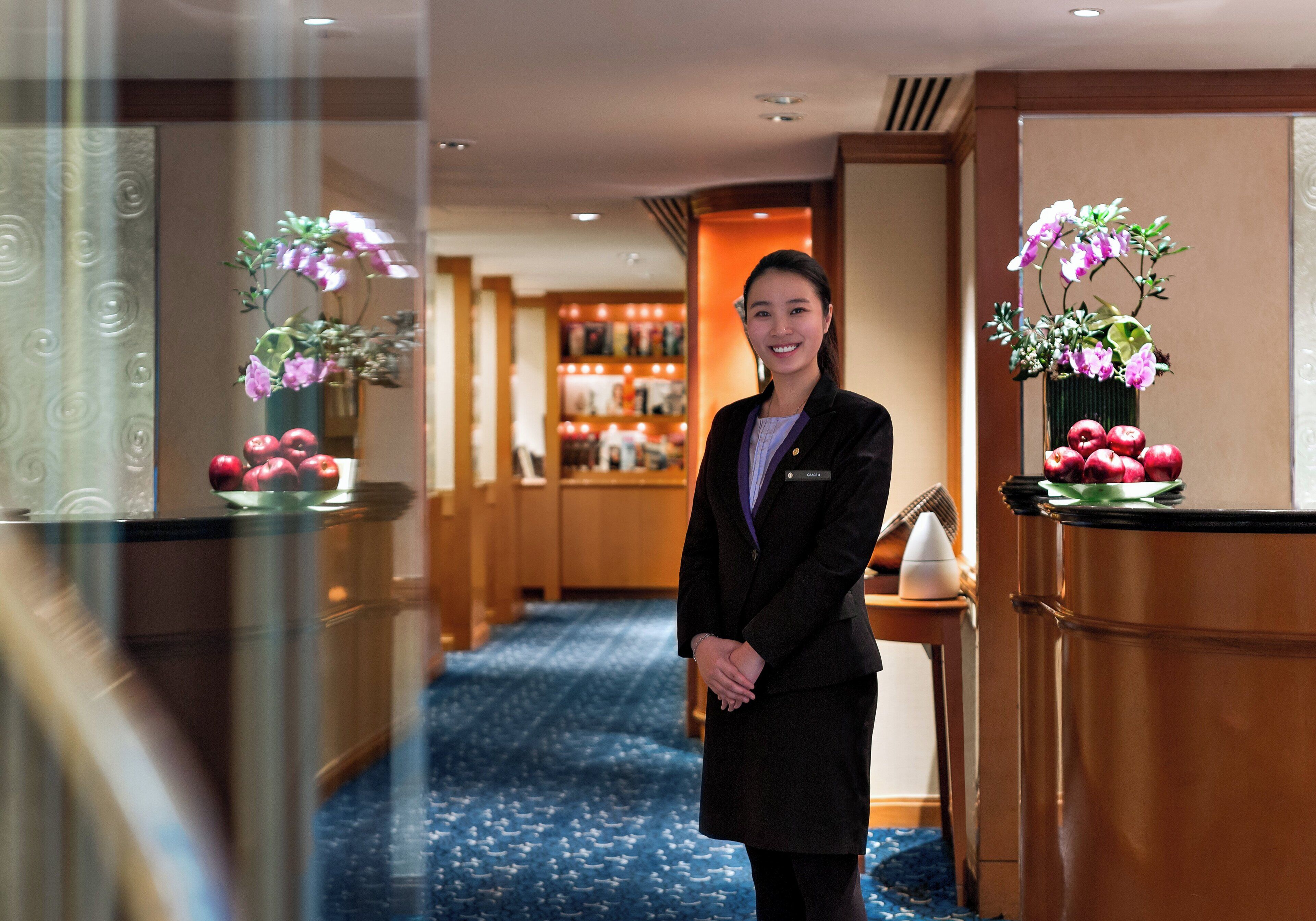 Um saguão de hotel com um balcão de recepção, piso coberto por carpete azul e uma exposição de flores e maçãs.