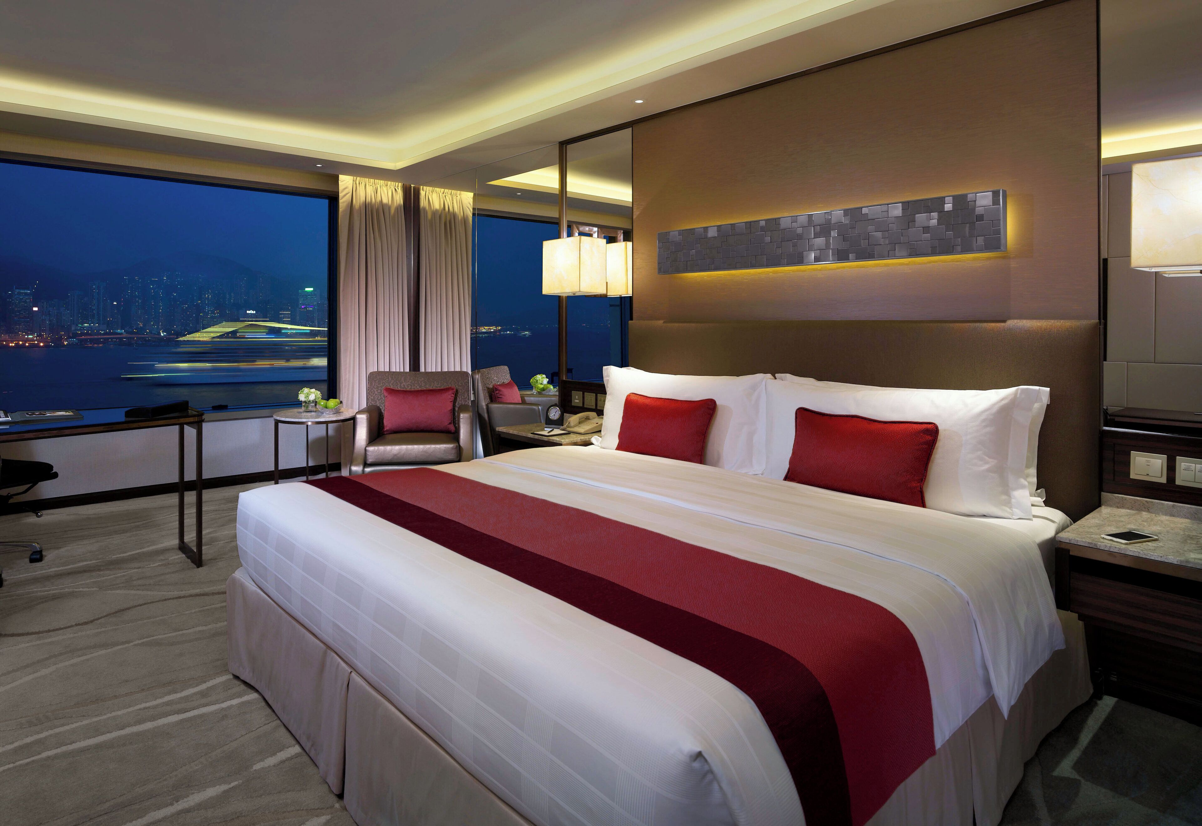 A hotel room with a large bed, a desk, a chair, a nightstand, and a view of the cityscape.