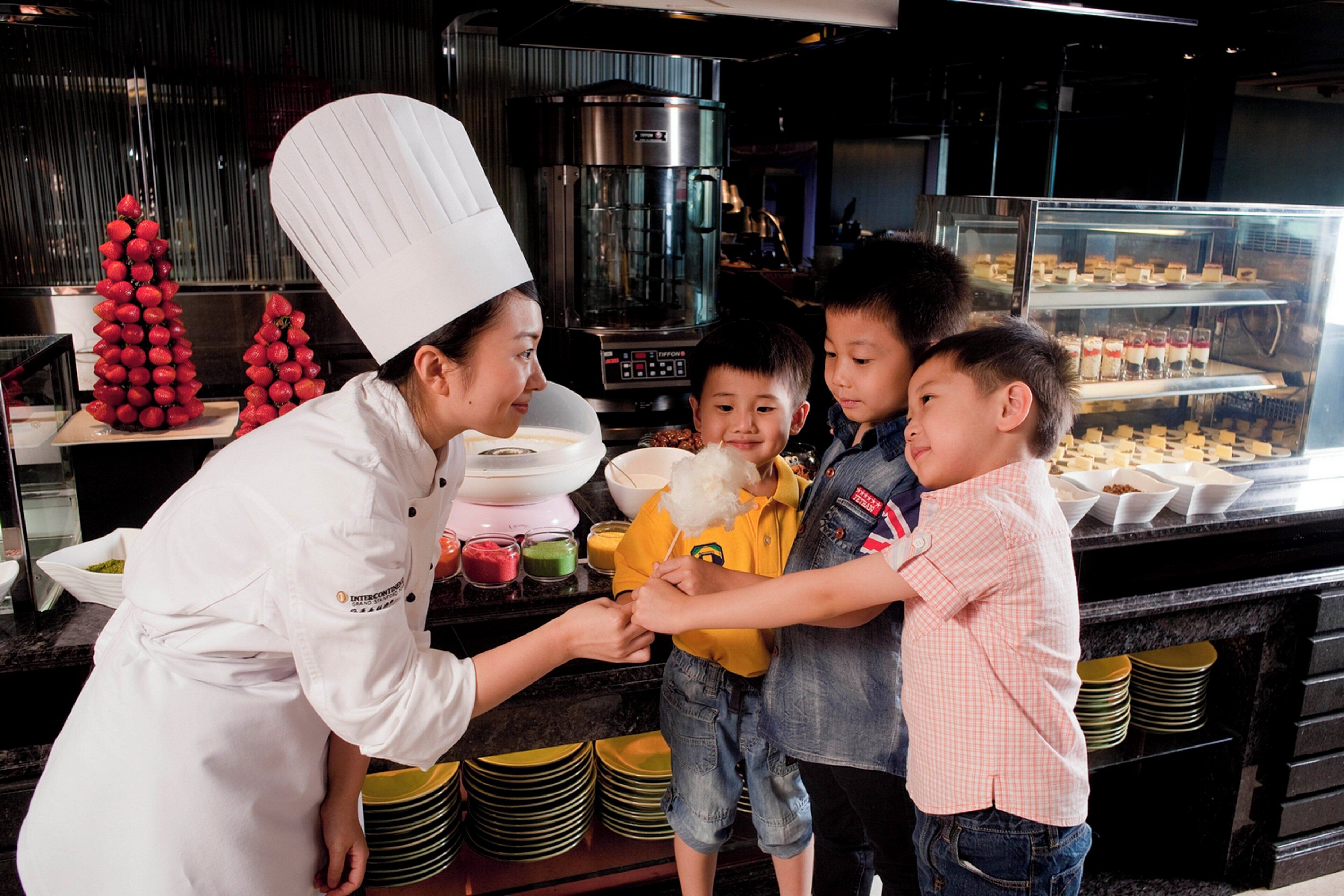 A chef interacting with children in a kitchen setting, with a coffee machine and dessert display in the background.