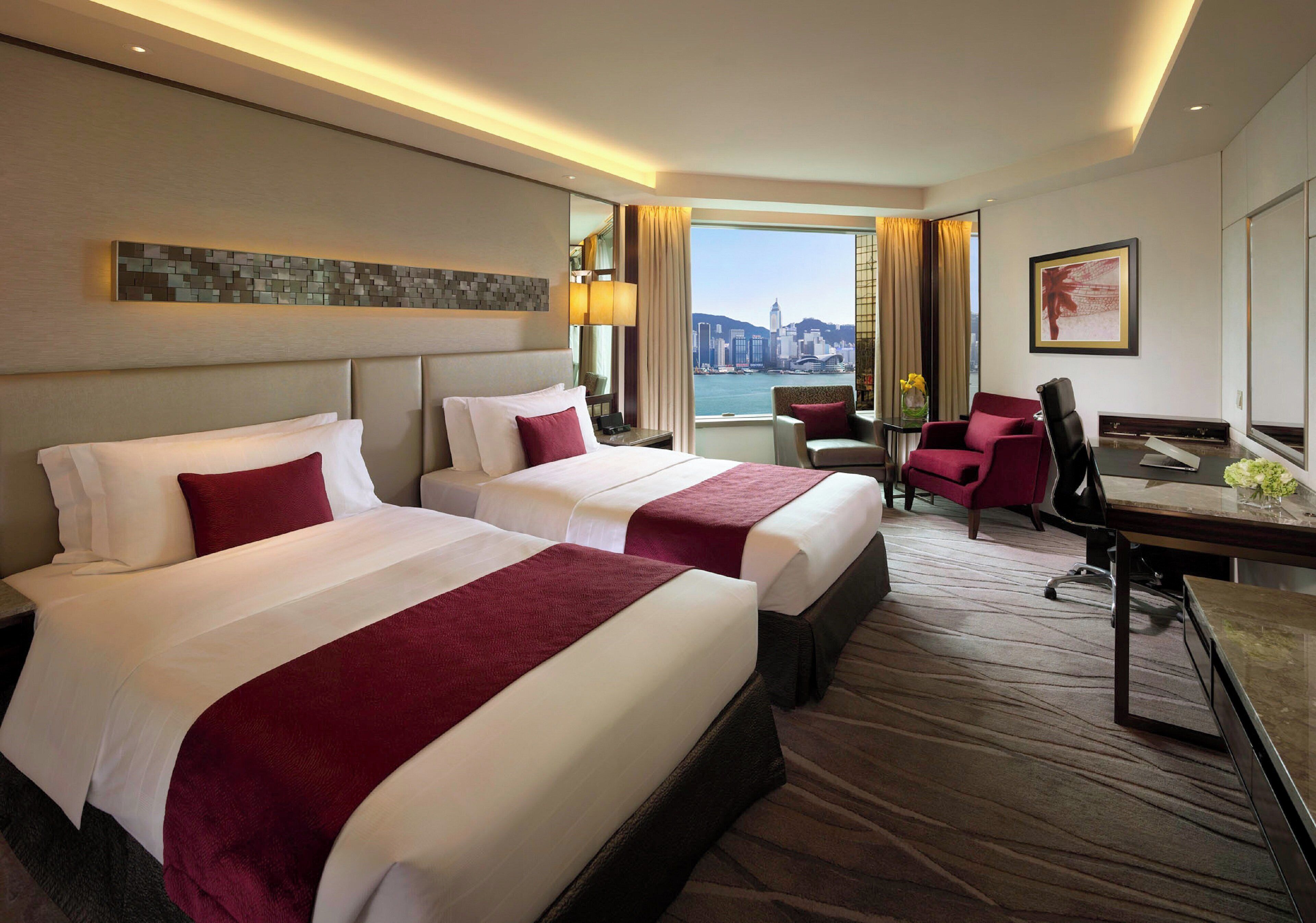 A hotel room with two beds, a desk, a chair, and a view of a cityscape.