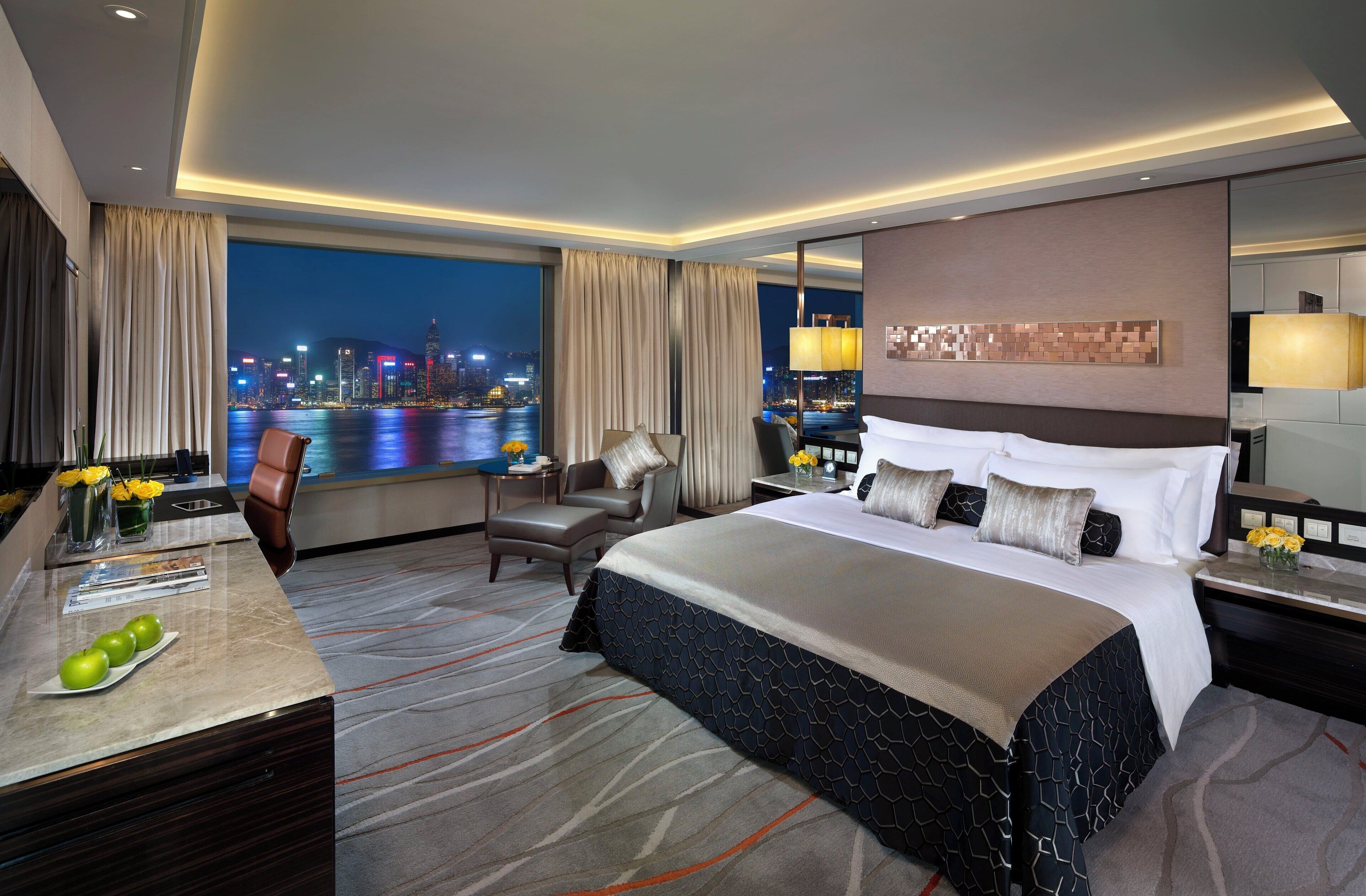 Une chambre d’hôtel avec un grand lit, un bureau avec une chaise, une vue sur la ville de nuit et un panorama de la ligne d’horizon urbaine par la fenêtre.