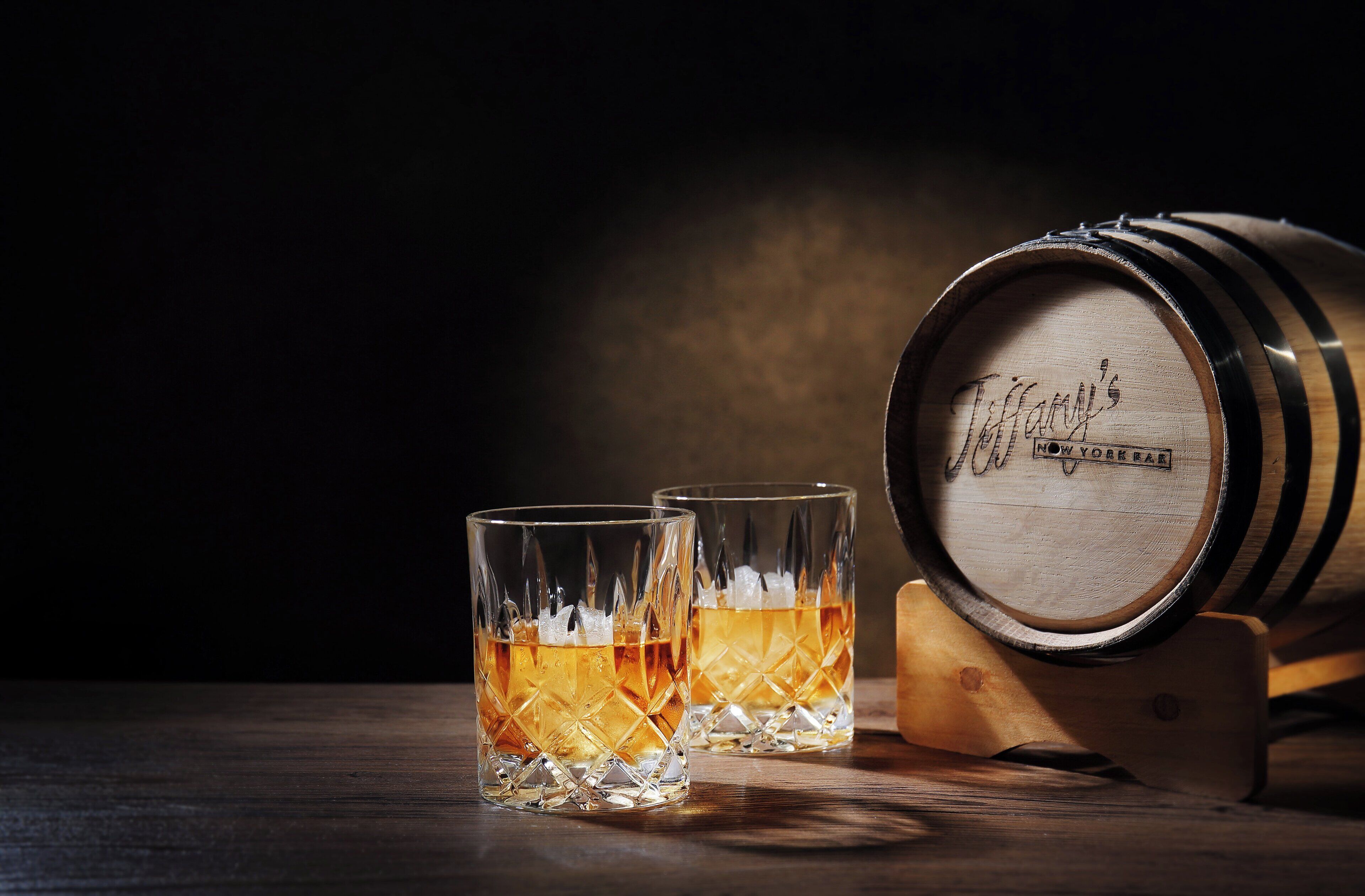 Two glasses of whiskey on a wooden table with a barrel in the background.