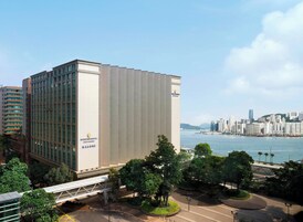 A modern hotel with a glass facade, located near a waterfront with a city skyline in the background.