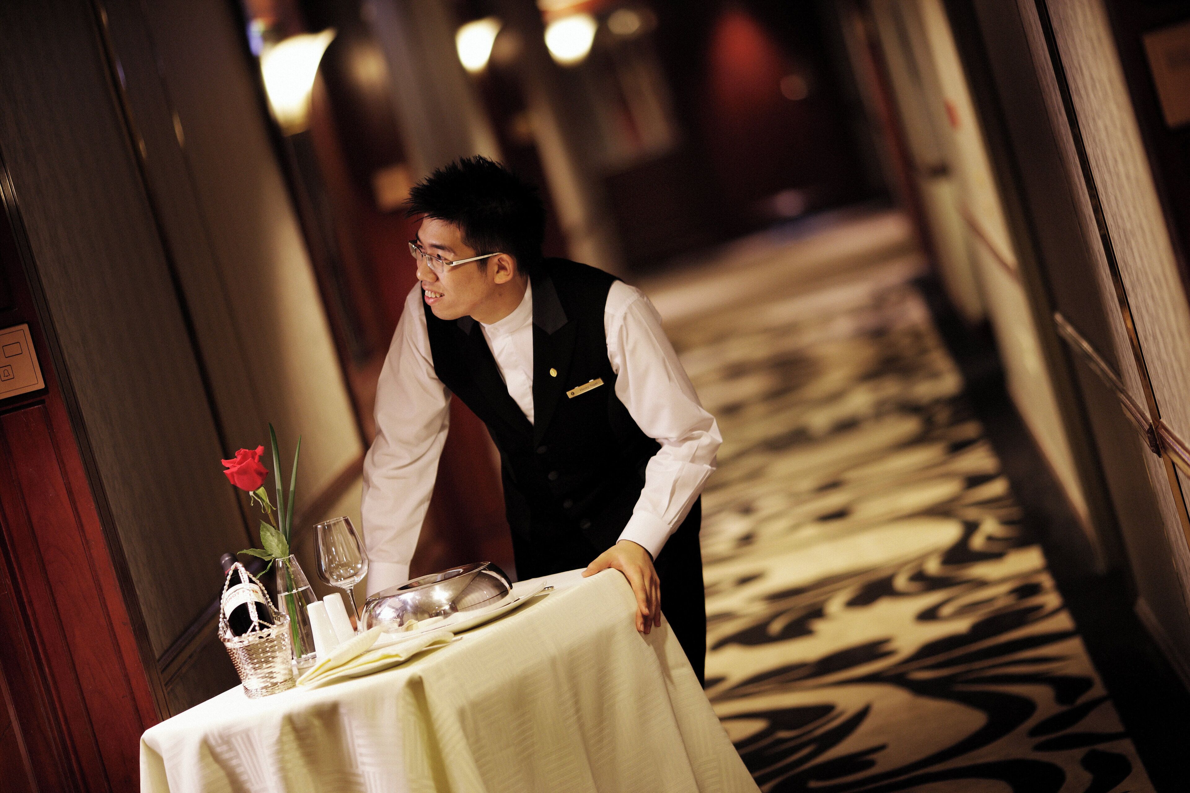Un couloir d’hôtel avec une table dressée pour le dîner, agrémentée d’un vase contenant une rose rouge, d’un verre à vin et d’un verre à eau.
