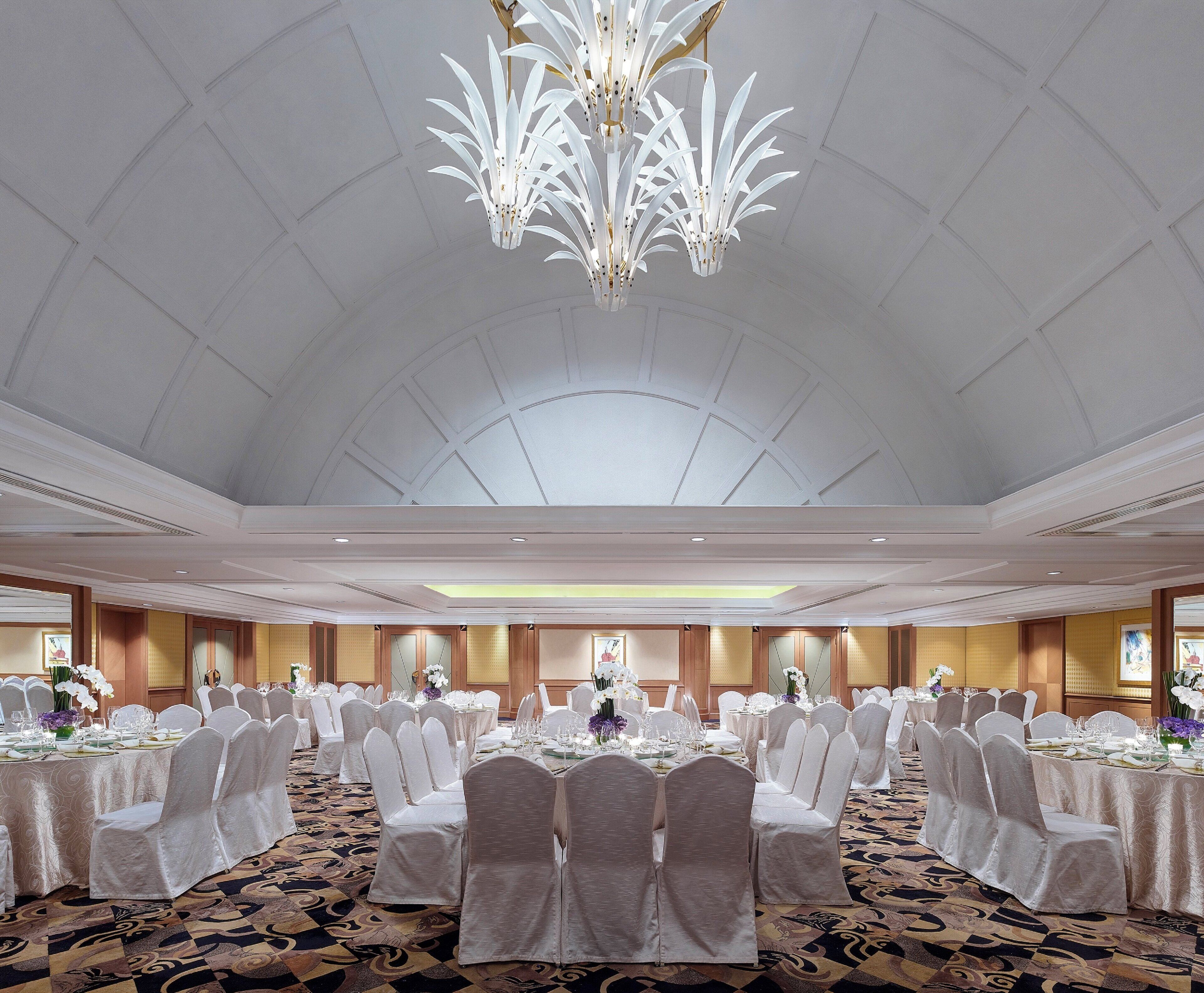 Un gran salón de banquetes con mesas redondas, sillas blancas y una lámpara central.