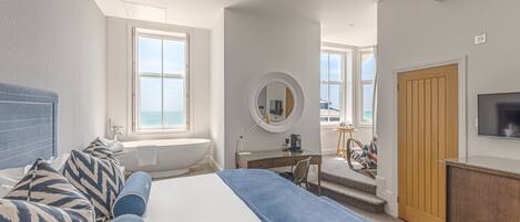 A modern hotel room with a large bed, a desk, a chair, a round mirror, a TV, and a view of the sea through large windows.