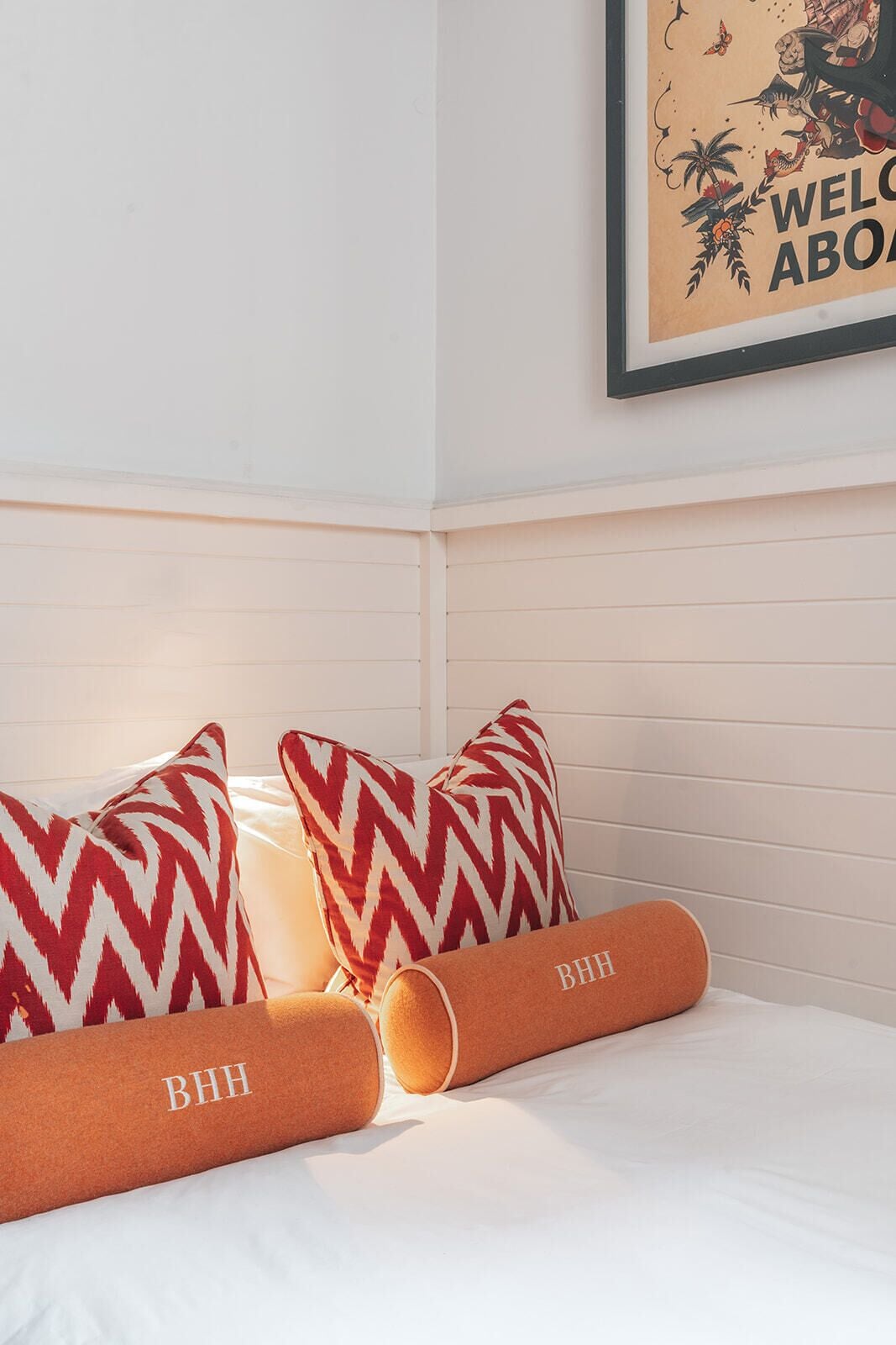 A hotel room with a bed, two patterned pillows, a rolled-up pillow with "BHH" embroidered, and a framed welcome sign on the wall.