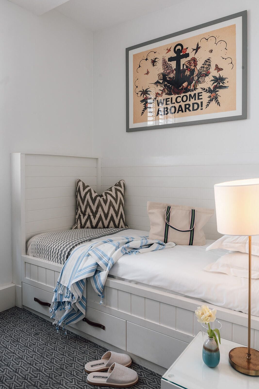 Un dormitorio con cama, almohadas, una mesita de noche con lámpara y un cartel de bienvenida enmarcado.