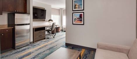 A modern hotel room with a kitchenette, a sofa, a desk, and two framed pictures on the wall.