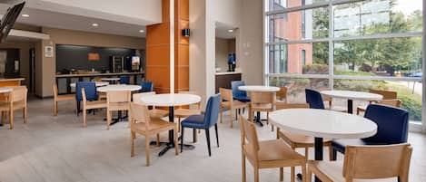 Een modern café met ronde tafels en stoelen, een keukenhoek en grote ramen met uitzicht op de omgeving.