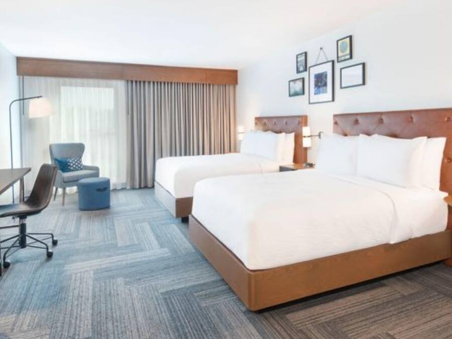 A hotel room with two beds, a desk, a chair, a lamp, and a large window with curtains.