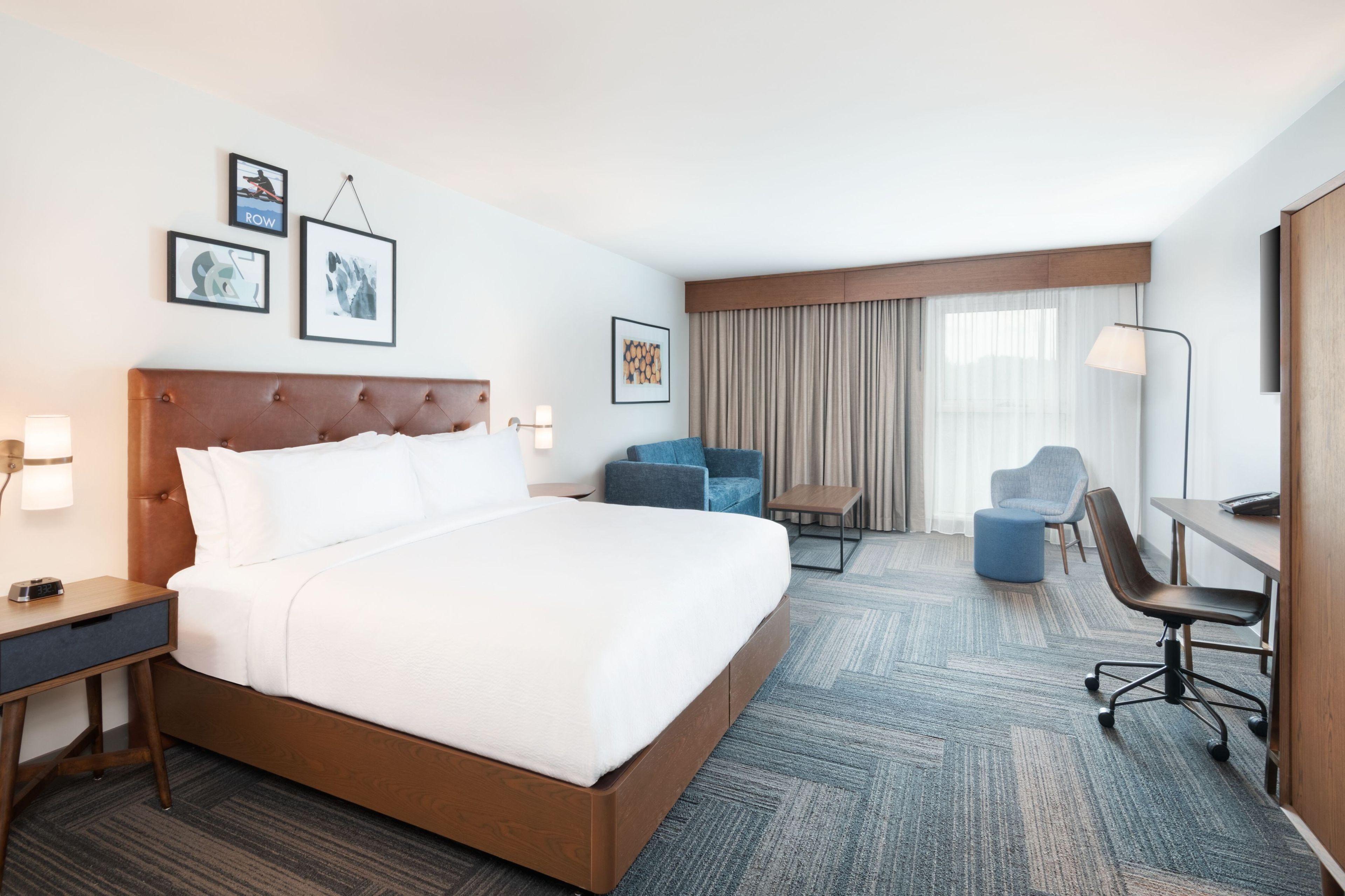 A hotel room with a large bed, a desk, a chair, and a sofa. There are framed pictures on the wall and a window with curtains.