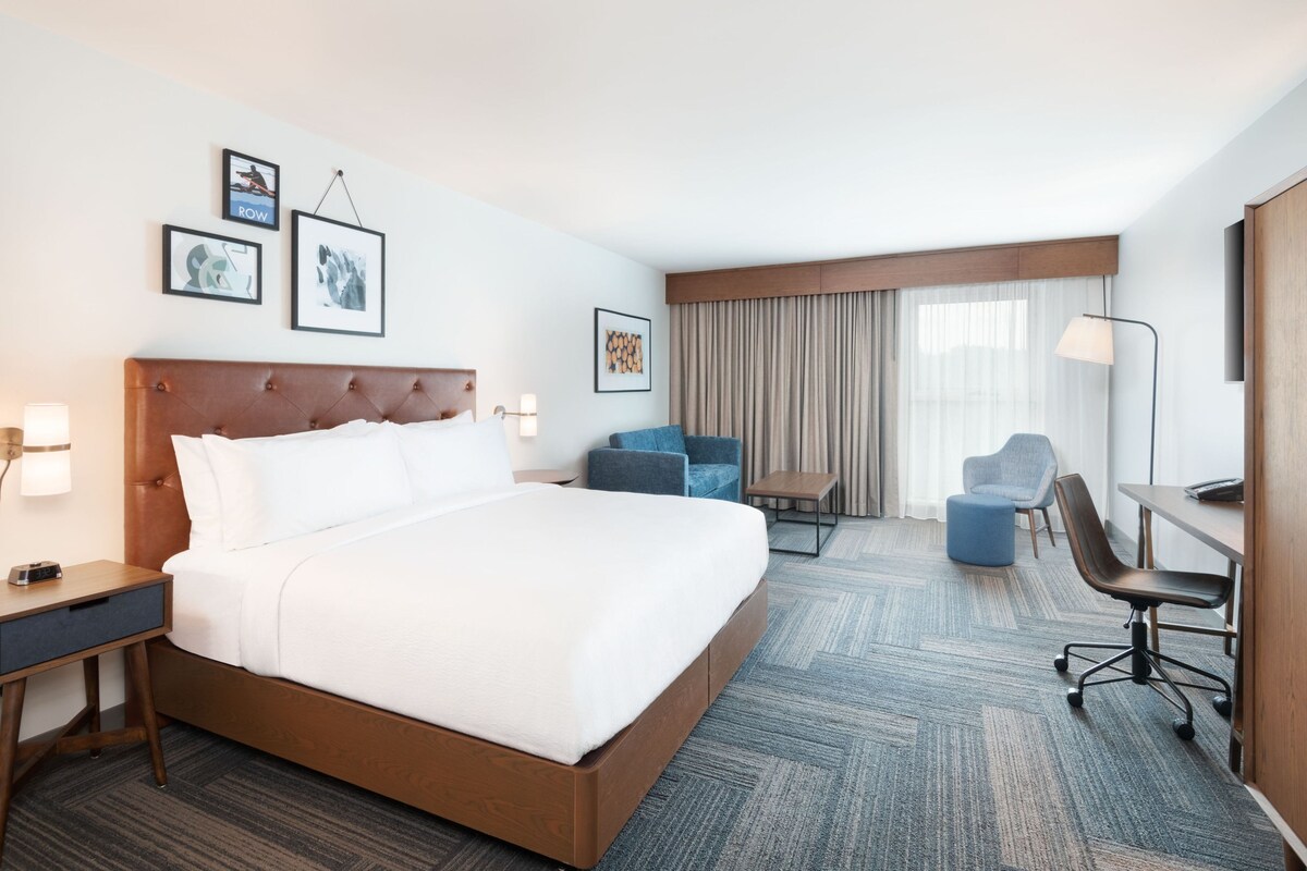 Habitación de hotel con una cama grande, un escritorio, una silla y una mesita. Hay cuadros en la pared y una ventana con cortinas.