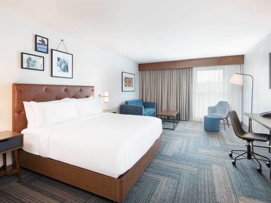 Quarto de hotel com uma cama grande, uma escrivaninha, uma cadeira e um sofá. Há quadros nas paredes e uma janela com cortinas.
