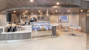 Una cafetería moderna con un expositor de vidrio, zona de estar con mesas y sillas redondas, y techo de madera.
