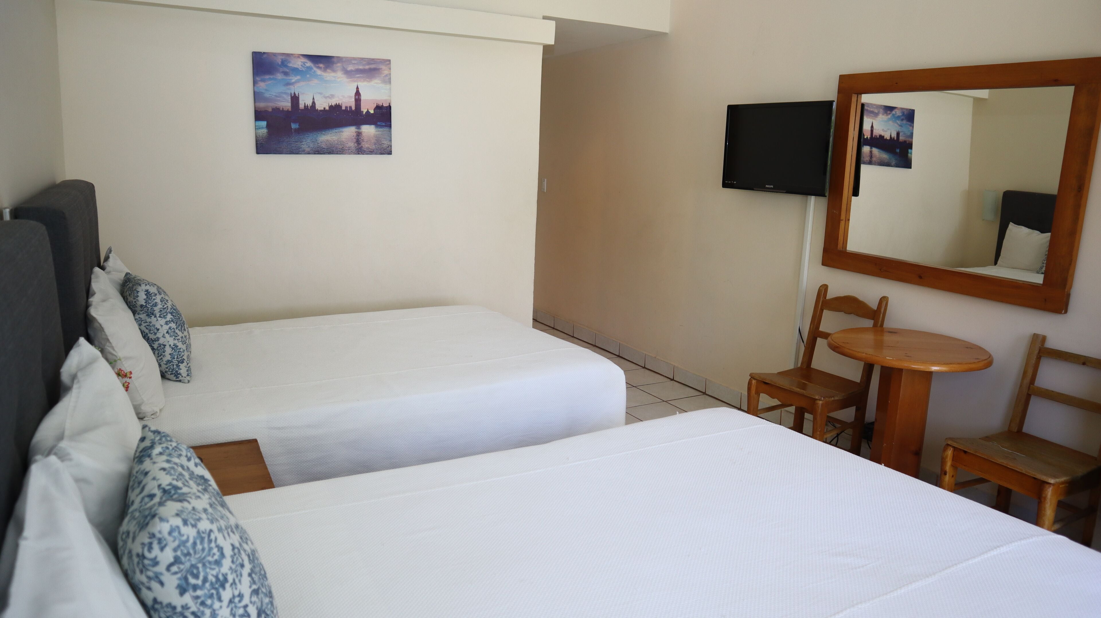 Hotellihuone, jossa on kaksi erillistä sänkyä, pieni puinen pöytä, tuoli, televisio ja seinällä kaupunkimaisemaa esittävä taulu.