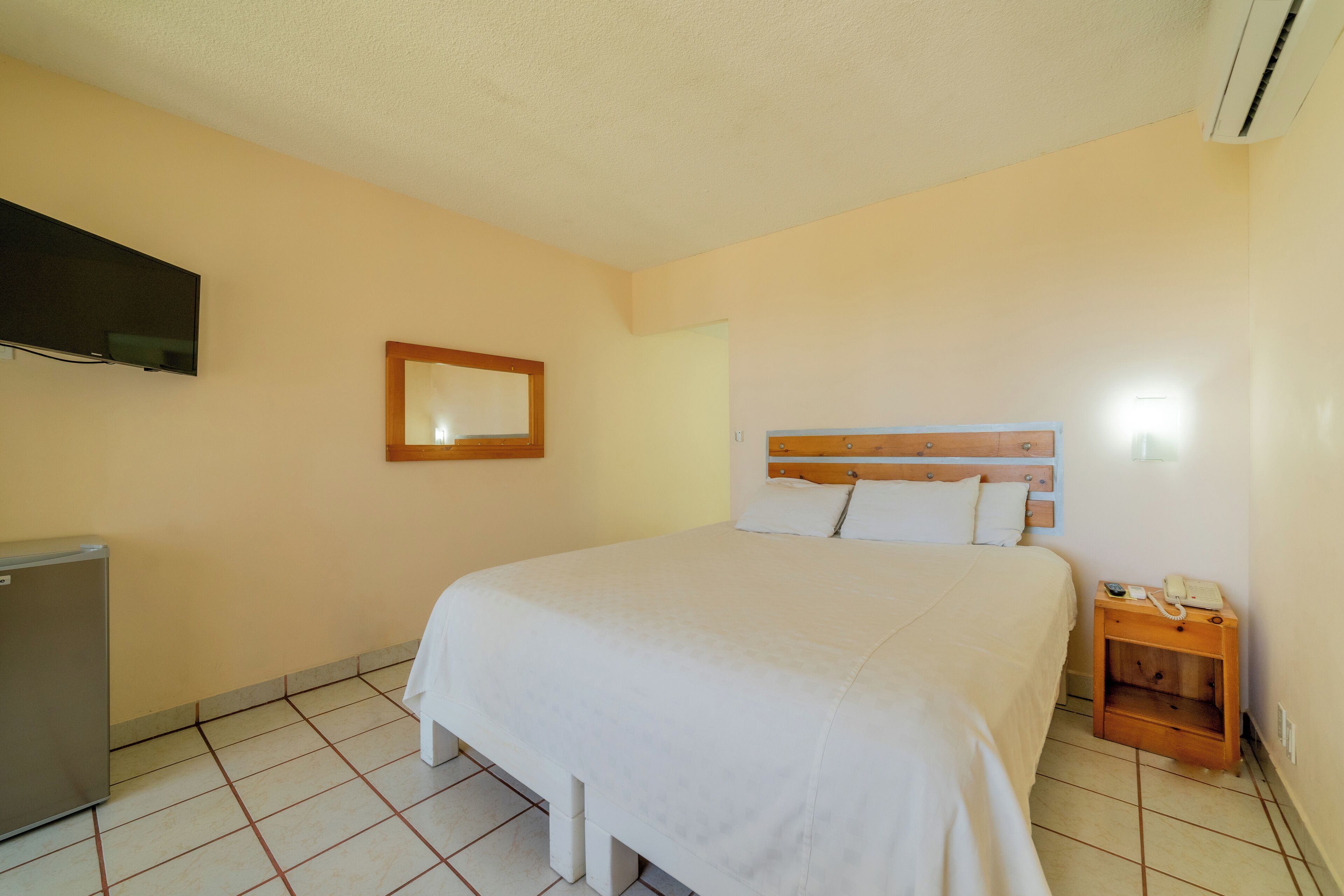Un dormitorio con cama, televisor montado en la pared, mesita de noche con lámpara y un refrigerador pequeño.