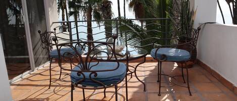 Una terraza con una mesa, dos sillas, un ventanal de vidrio y vistas a palmeras y al mar.