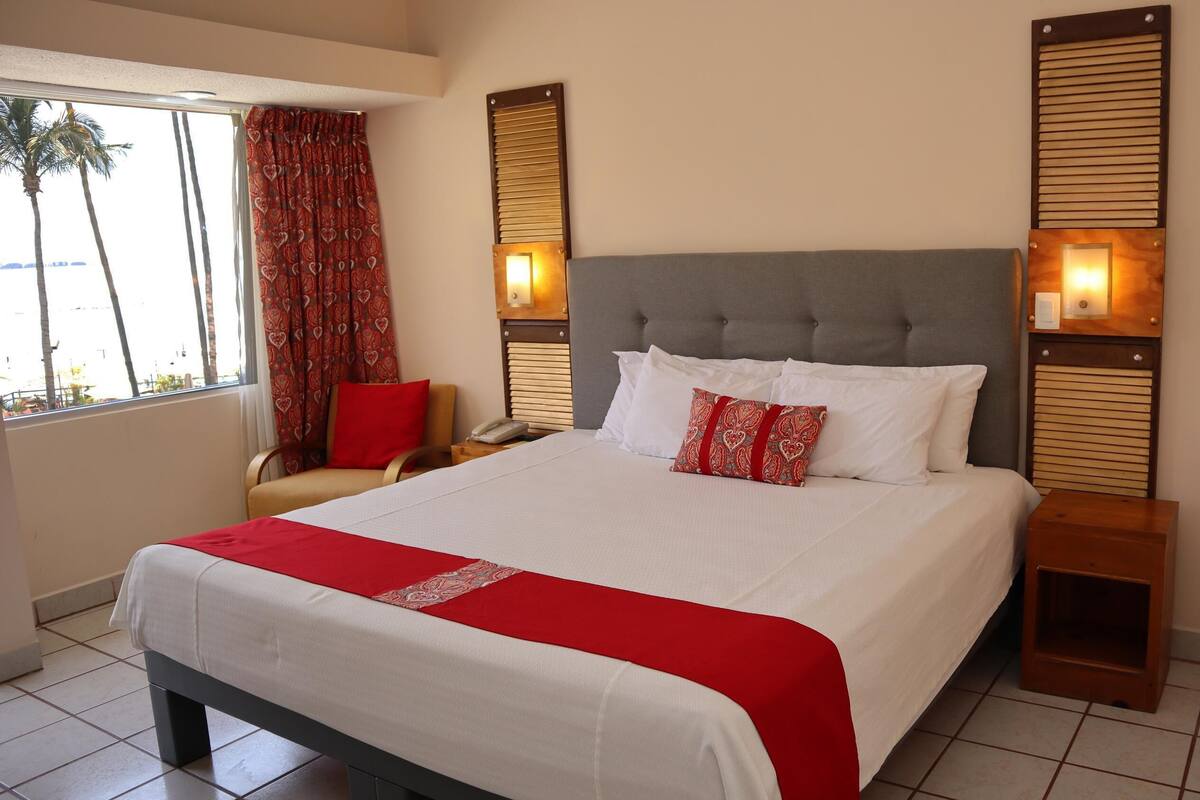 A hotel room with a large bed, a chair, a bedside table, and a window with curtains.