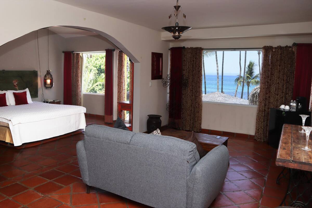 A hotel room with a bed, a sofa, a dining table, and a view of the ocean.