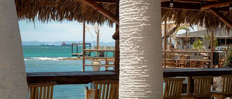 A beachside restaurant with wooden chairs and a thatched roof.