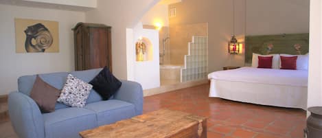 A room with a blue sofa, a wooden chest, a bed with red pillows, a shower, and a staircase.