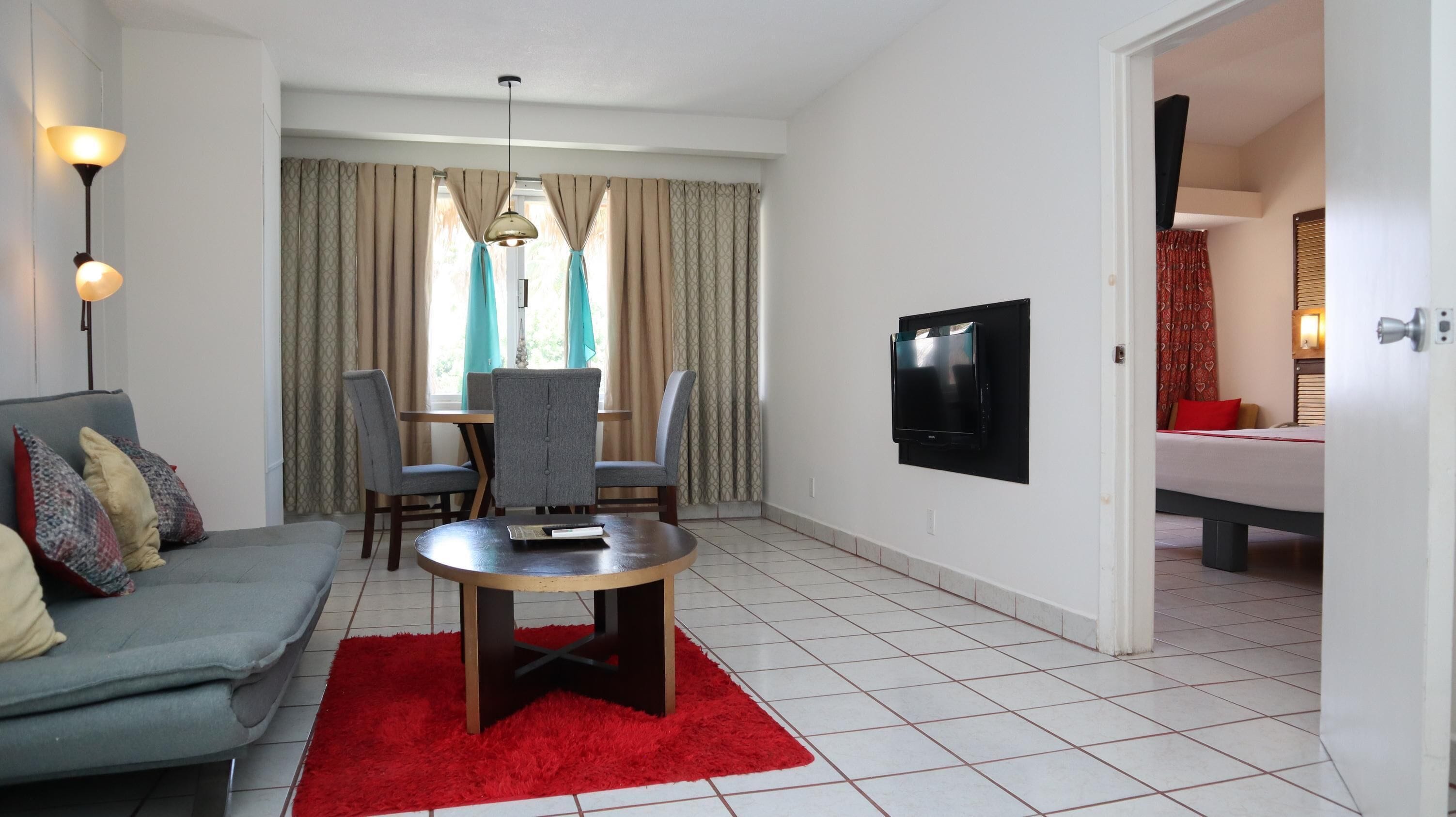 A modern hotel room with a grey sofa, a red rug, a dining table with chairs, a TV mounted on the wall, and a bed with a red pillow.